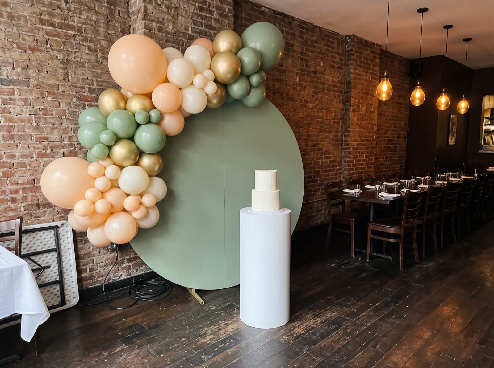Round sage backdrop with a peach and ivory balloon garland and a cake on a pedestal in a restaurant private dining room